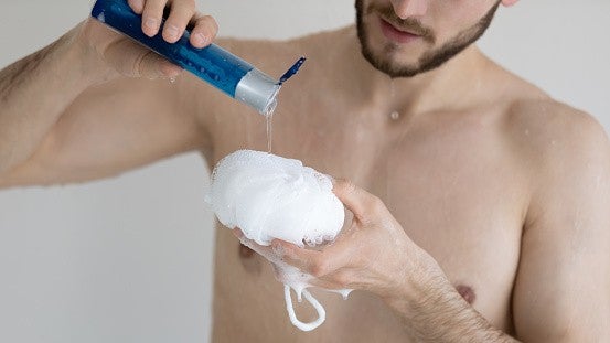 Close up young man applying moisturizing gel liquid soap on fluffy bath puff, preparing to taking shower, washing body, standing in bathroom at home, skincare and personal hygiene concept