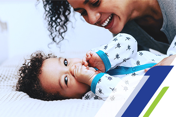 Uma mulher sorrindo e cuidando de seu neném que está deitado na cama com as mãos na boca
